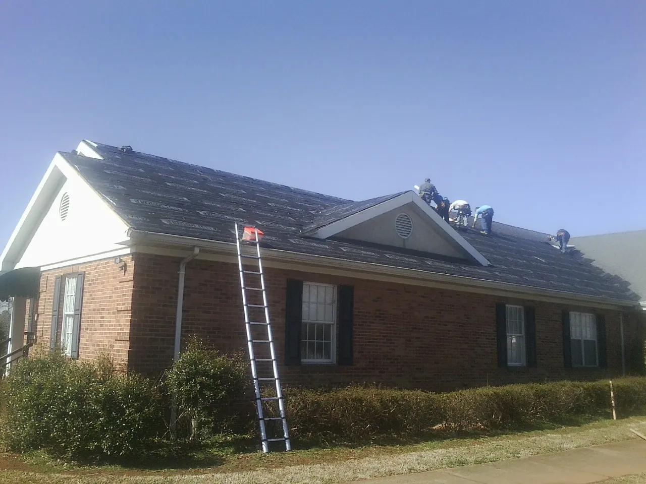 Crew tearing shingles off a brick ranch roof with a ladder set — Emergency Roof Repair work by our crew in Fontana