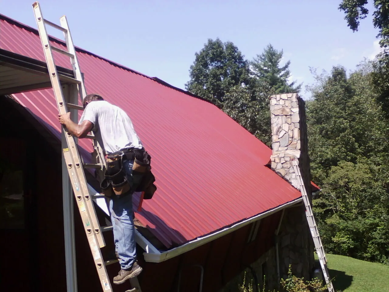Roofer on a ladder beside a red metal roof and stone chimney — Soffit & Fascia Repair by Roofing Brothers in Fontana